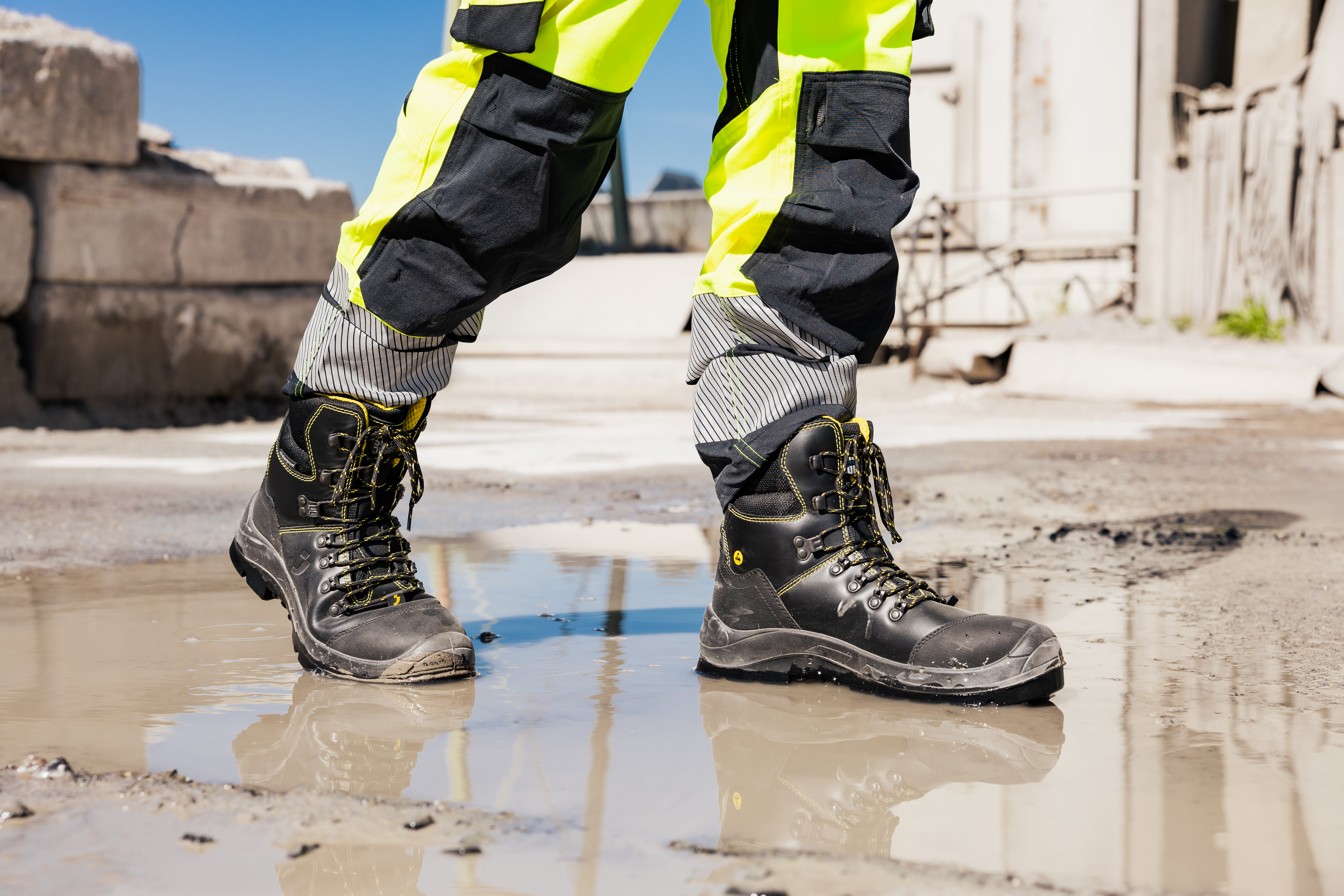 Worker in high-visibility trousers and black safety boots walking through a puddle on a wet construction site.